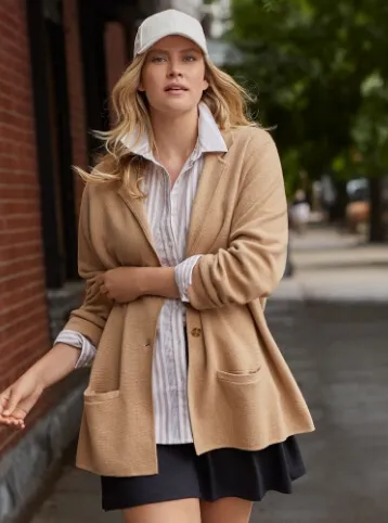 Woman wearing a white dress shirt under a tan cardigan, with a black skirt and white ball cap; walking down an urban sidewalk.