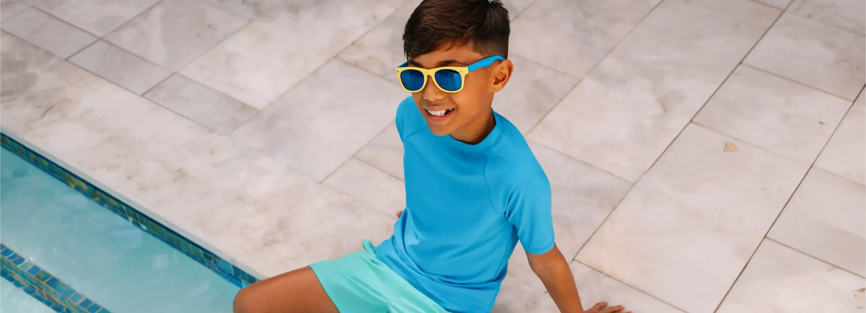 Boy wearing a blue rash guard, swim trunks and sunglasses sitting poolside.