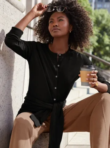 Woman wearing a black lightweight cardigan and tan pants; holding a coffee cup while lifting her sunglasses to gaze into the distance.