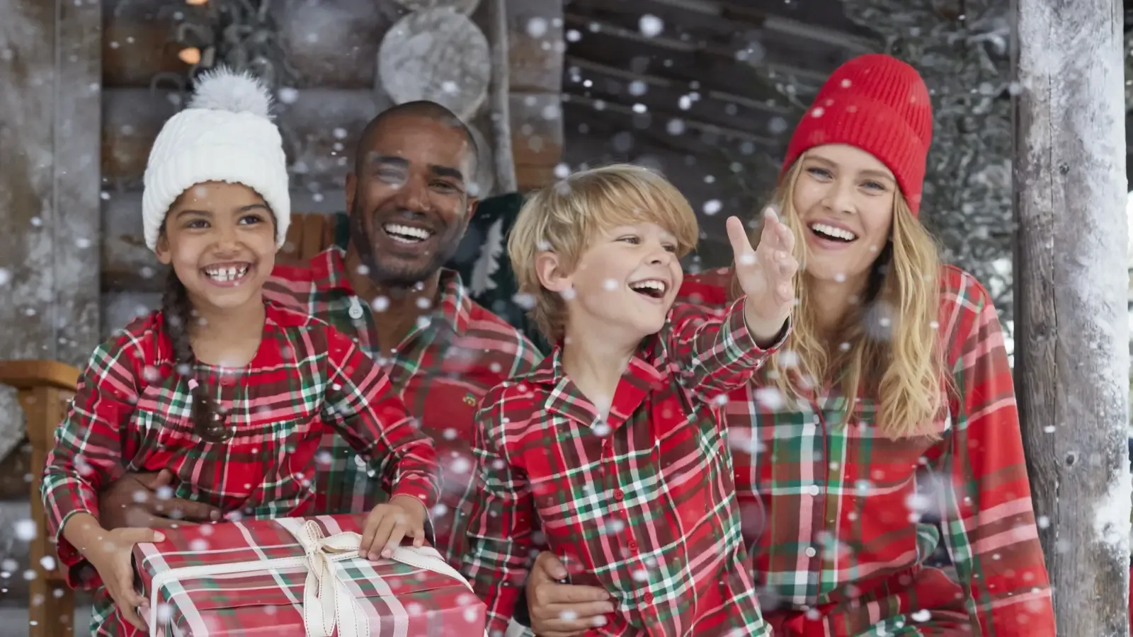 A family wearing matching red plaid pajamas poses for a picture on the snowy porch of a log cabin.