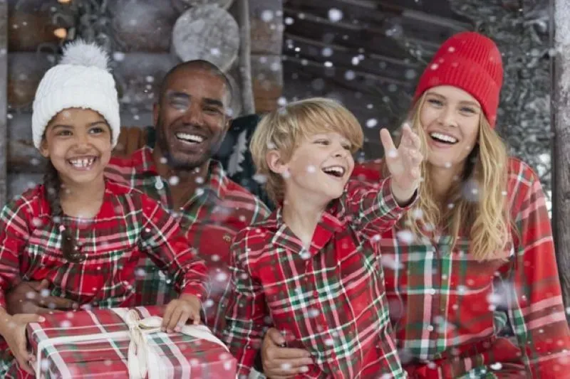 A family wearing matching red plaid pajamas poses for a picture on the snowy porch of a log cabin.