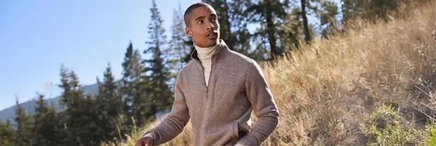 Man wearing a gray quarter zip fleece over a white turtleneck; standing on a grassy hillside.