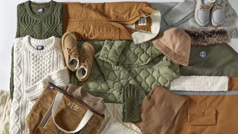 Several green, brown and beige sweaters, vests, jackets, boots and accessories laid out neatly.