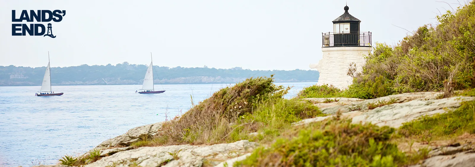 Lighthouse on a rocky shoreline that's covered with vegetation and moss; two sailboats going by in the middle distance.