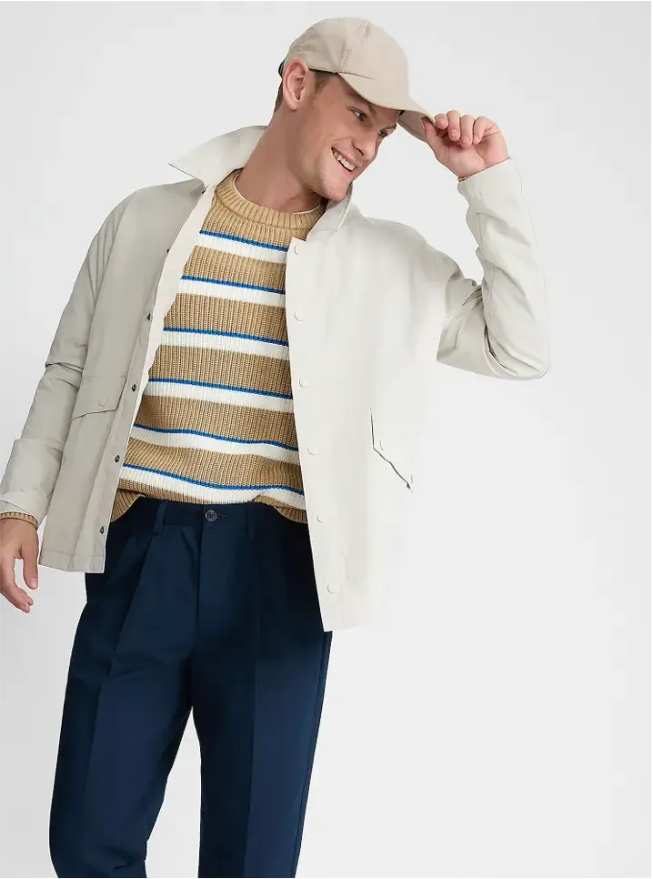 Man in a cream jacket over a beige and blue striped sweater with navy trousers.