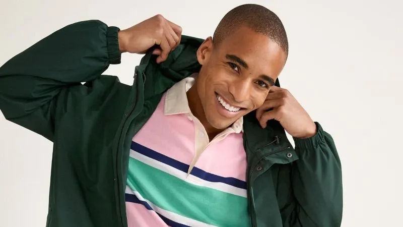 A man lifts the hood of a green rain jacket worn over a striped rugby shirt.