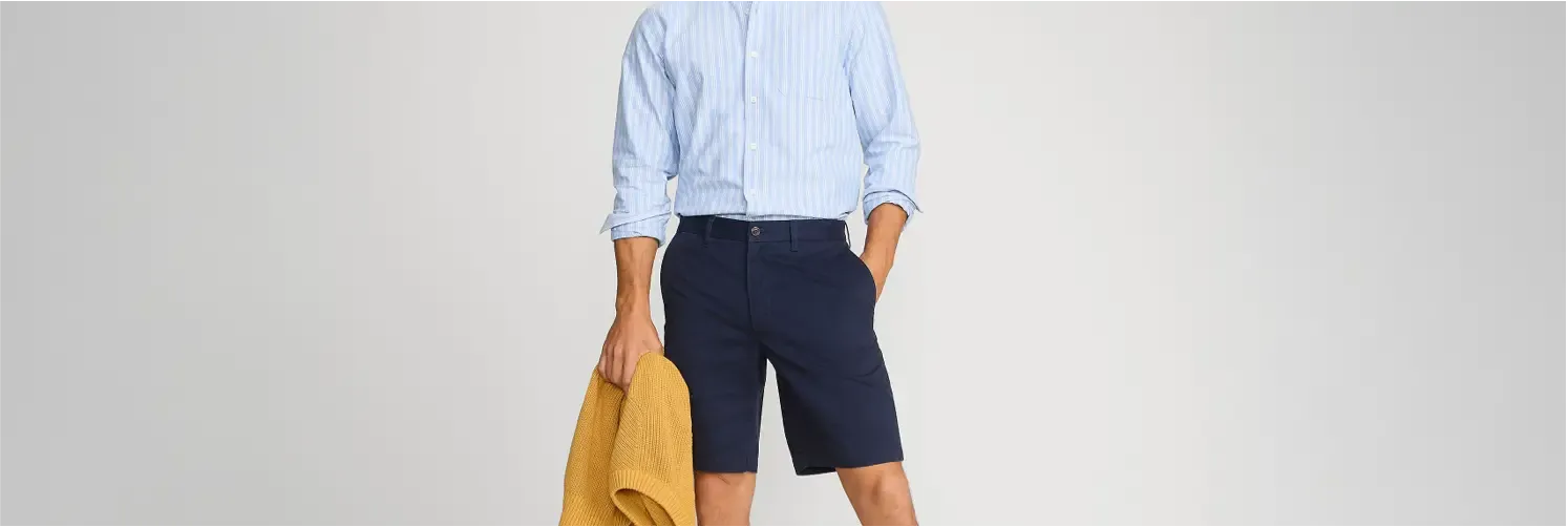 Man wearing a light blue striped button-up shirt and navy shorts, holding a mustard sweater.