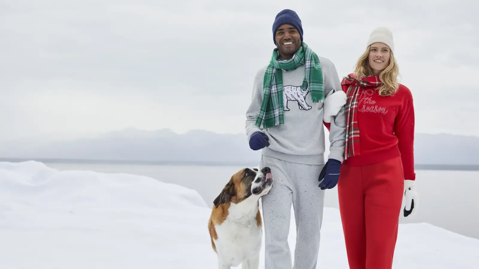 A man and woman dressed in holiday sweat suits, hats, gloves and scarves, walking their dog outdoors in a snowy landscape.