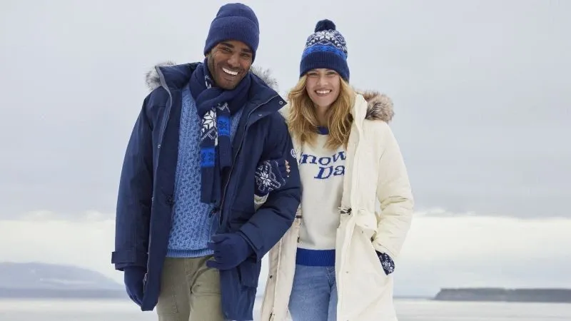 A man wearing a navy parka and woman wearing a white parka over seasonal sweaters, both wearing a winter hat and gloves.
