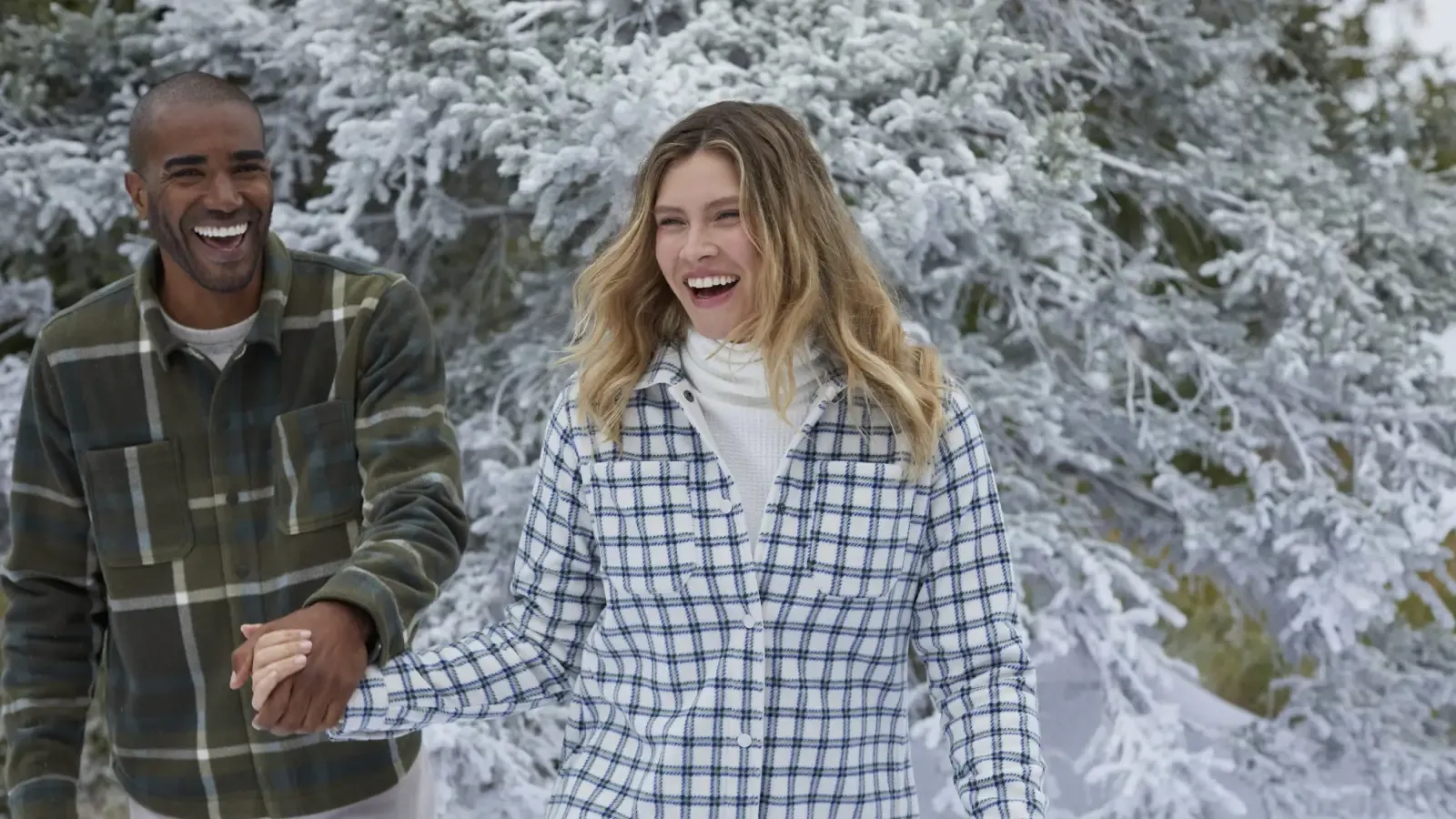 A man and woman wearing plaid fleece shirt jackets hold hands near snow covered evergreen trees.