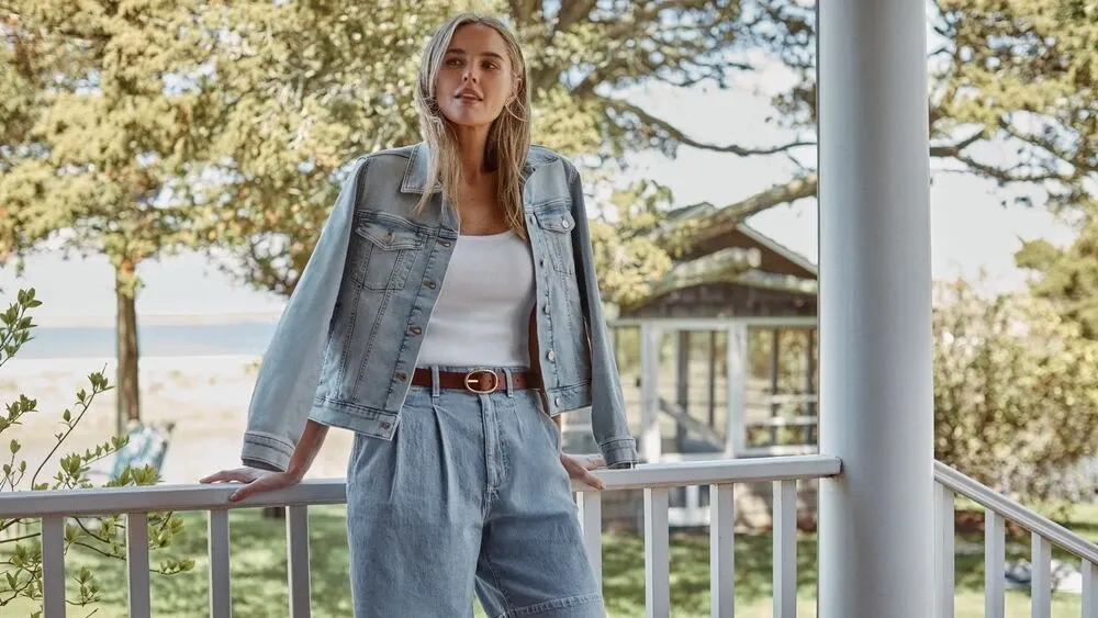 Woman wearing a white shirt and jean shorts with a denim jacket over her shoulders.