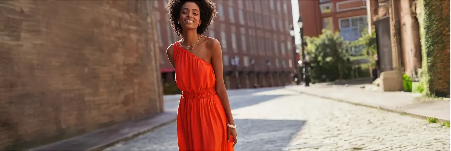 Woman wearing a bright red-orange one-shoulder midi dress walking down an urban street.