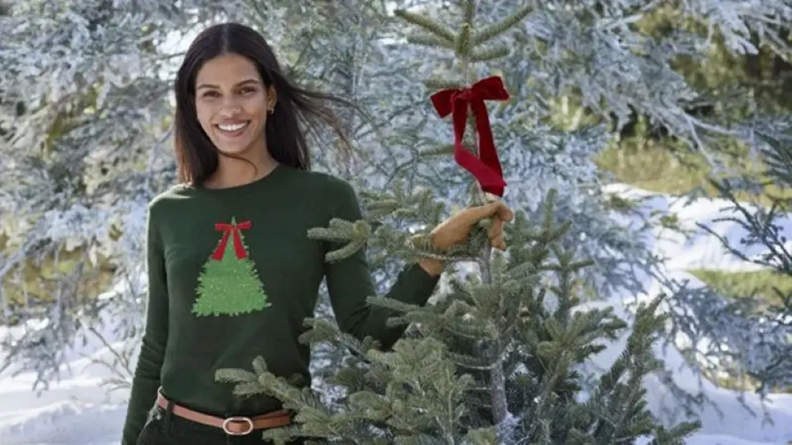 A woman wearing a green sweater with a Christmas tree and bow, standing next to a small pine tree with a bow.