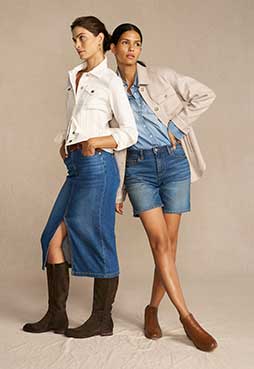 Two women pose with each other. One is wearing denim skirt and a white shirt. The other is wearing a shirt jacket with denim shorts.