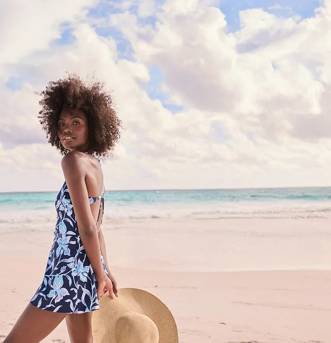 A woman wearing a floral pattern swim dress holding a wide brim sun hat.