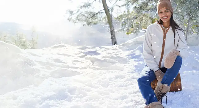A woman sitting in the snow wearing a tan beanie, a white coat, mittens, and blue jeans.