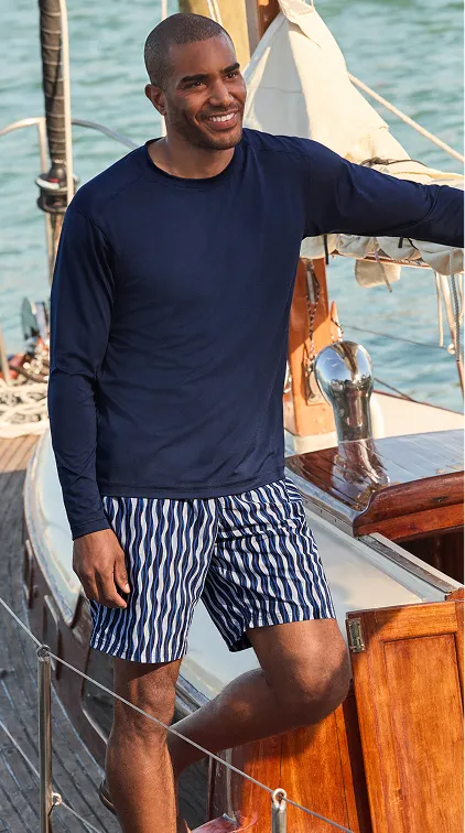 A man wearing a long sleeve navy top over white and navy striped shorts stands on the deck of a sailboat.