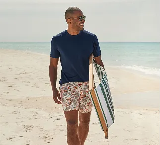 A man wearing a navy top and floral pattern swim trunks carries a chair on a beach.