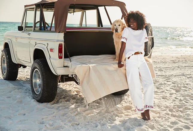A woman wearing white denim sits on the back of a jeep with her golden retreiver at the beach.