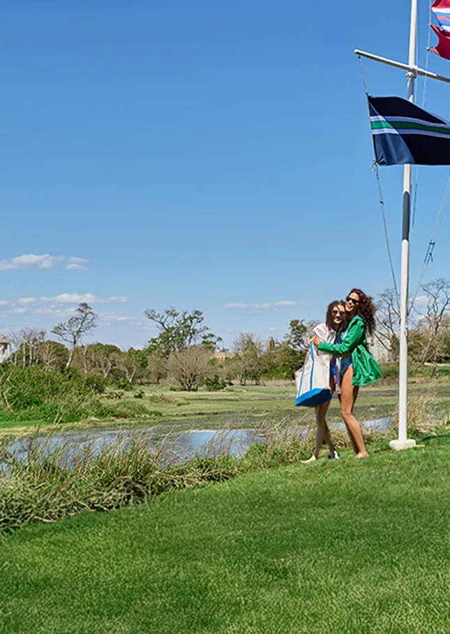 Two women standing by a flag pole with three flags on it next to a lake.