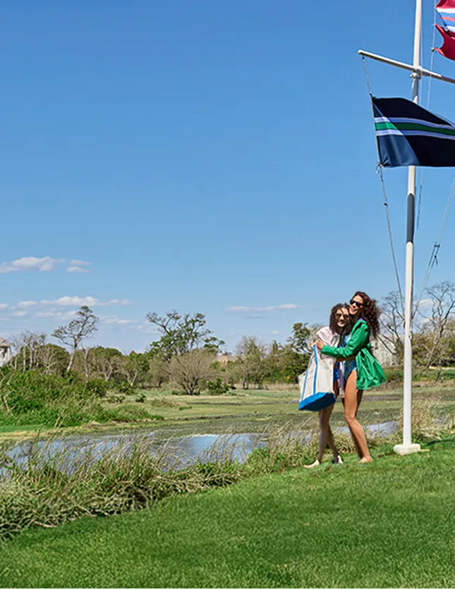 Two women standing by a flag pole with three flags on it next to a lake.