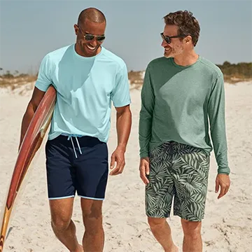 Two men walking on a beach in t-shirts and swim trunks.