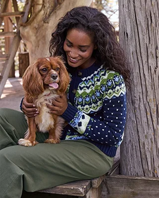 A woamn wearing a novelty holiday sweater holds a small dog that has fluffy ears.