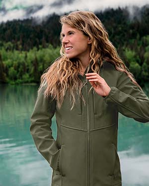 A woman wearing a olive jacket in front of a lake by a mountain.